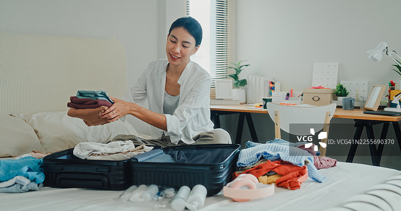年轻的亚洲女性正在卧室的床上打包行李箱，为轻松的夏日度假旅行准备衣物和个人物品。图片素材
