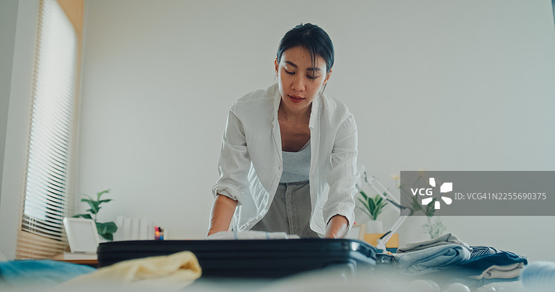 一位年轻亚洲女性在家里的卧室，正坐在床上整理行李，为即将到来的轻松夏日度假旅行准备衣物和个人物品。图片素材