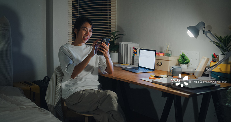 年轻亚洲女性在家中卧室的夜晚，使用智能手机开关灯，坐在床上。智能家居技术在日常生活中的应用概念。图片素材