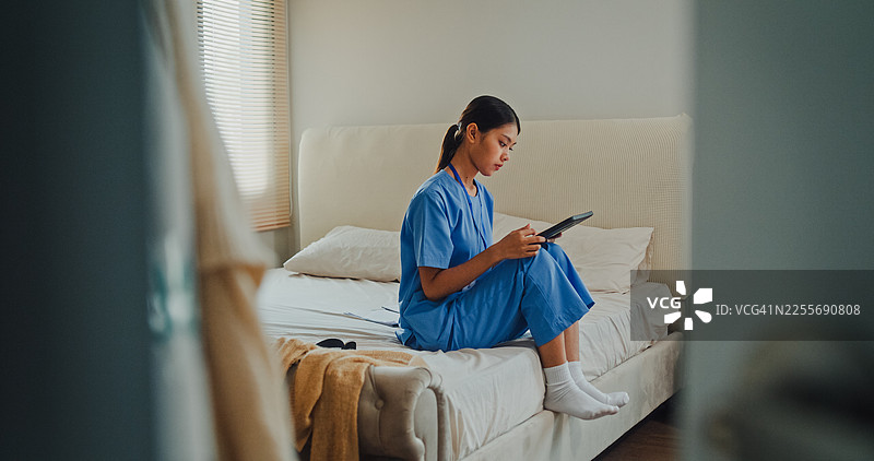 身穿蓝色工作服的年轻亚裔女护士坐在床边使用数码平板电脑。护士生活方式概念。图片素材