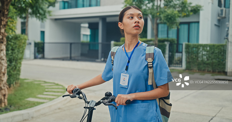 年轻的亚裔女护士穿着蓝色工作服，在宁静的居民区旁边骑着自行车。护士可持续生活方式的概念。图片素材