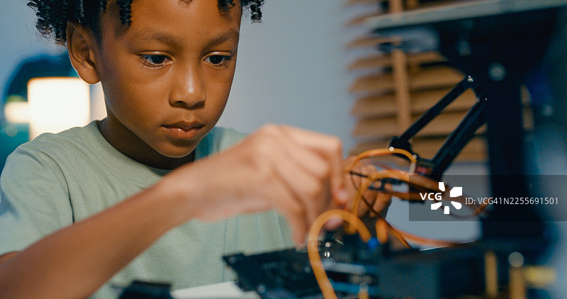特写：一个年轻的非裔美国男孩在家中夜晚仔细组装机器人汽车，展示他在科学和技术方面的技能。STEM教育机器人在线课程。图片素材
