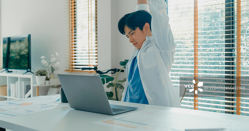 年轻的亚洲男性在家工作时休息伸展身体，强调人体工程学和放松的重要性。远程工作与金融。图片素材