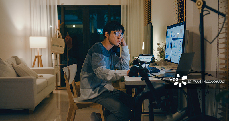 疲惫的年轻亚裔男员工在家中办公室加班，因多屏幕上的财务规划和数据分析任务而感到精神倦怠和不堪重负。远程办公与金融。图片素材