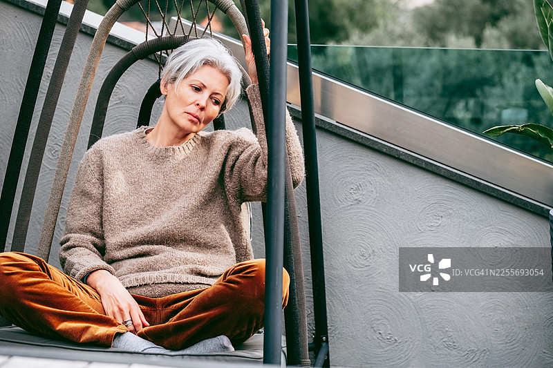 一位头发灰白的中年女性坐在户外的秋千椅上，一只手放在头边，若有所思或显得有些悲伤。图片素材