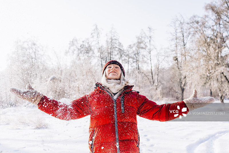 一位穿着红色冬装的快乐女子在雪地公园里伸开双臂，享受着雪花飘落。冬季乐趣，户外生活方式图片素材