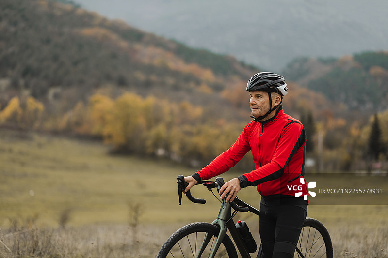 身穿红色夹克的年长骑行者，骑着碎石自行车，在秋季乡村景色中休息并欣赏风景图片素材