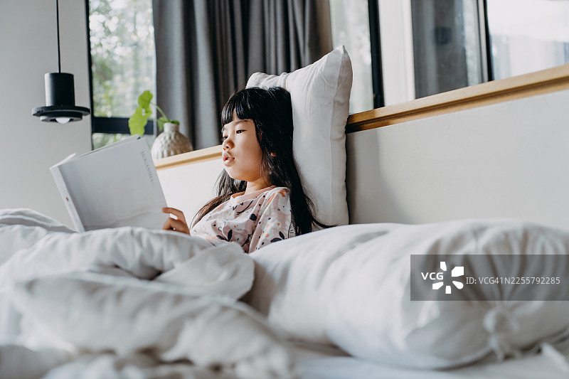 亚洲女孩早晨在卧室的床上放松地看书，窗户洒进自然阳光。儿童学习、教育和语言发展概念图片素材