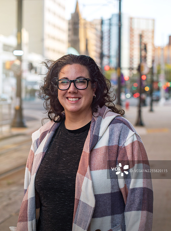 新泽西州纽瓦克市，一名深色头发的白人女性对着镜头微笑的生活方式肖像照，背景是模糊的城市建筑和交通信号灯。图片素材