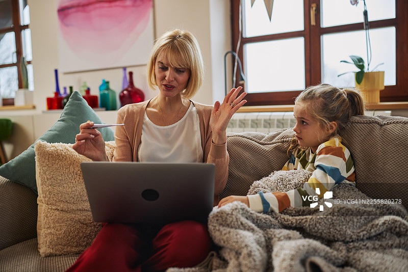 温馨居家关怀时刻：母亲正在与医生进行视频通话，生病的孩子则在沙发上休息图片素材