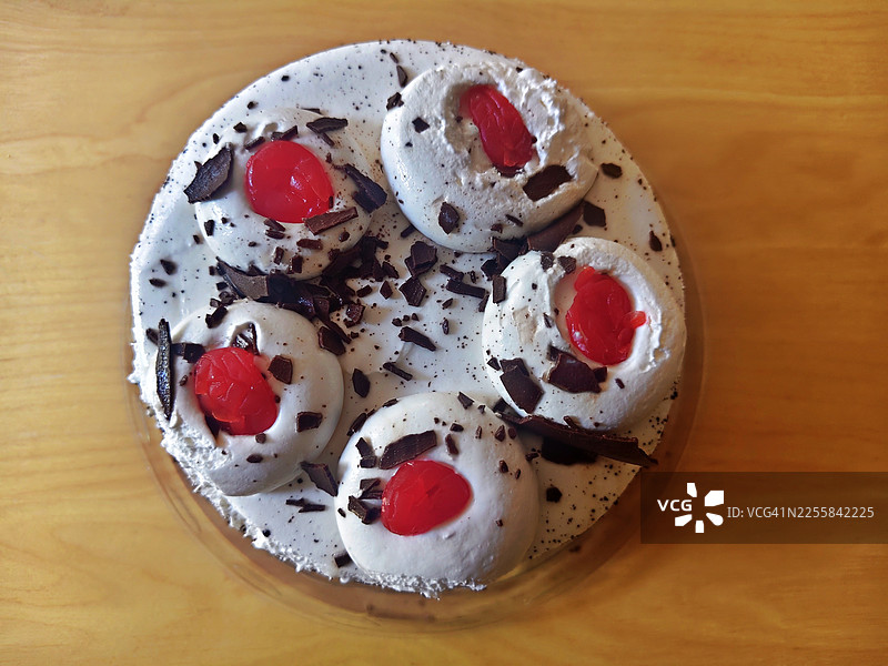 一个经典的黑森林蛋糕，上面点缀着奶油，每个奶油上放着一颗樱桃，并撒上了巧克力碎屑，放在陶瓷盘子里，盘子置于木桌上图片素材