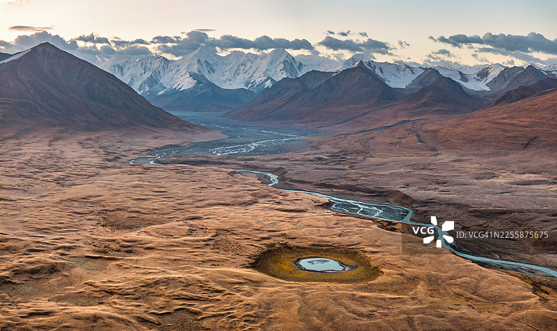 黎明时分，吉尔吉斯斯坦天山山脉的红棕色高地高原上，蜿蜒的冰川融水和一个独特的圆形黑湖占据了主导地位。图片素材