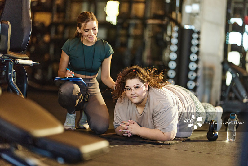 一位微笑的女教练在健身房指导一位肥胖女性进行体育训练。图片素材