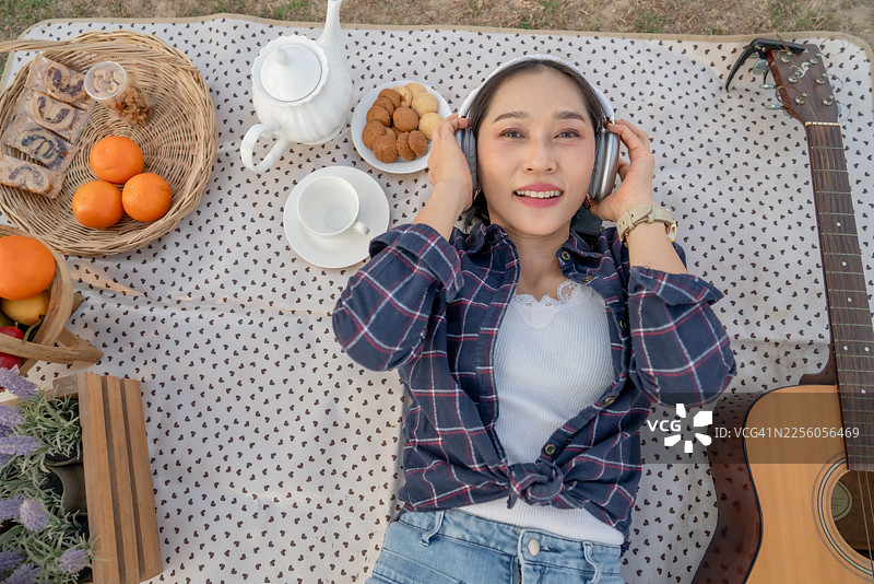从上方视角看，一位快乐的年轻亚洲女性躺在公园的野餐垫上，戴着耳机听音乐，旁边有原声吉他和食物，享受着悠闲的夏日周末生活。图片素材
