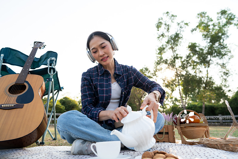 夕阳西下，一位快乐的年轻亚洲女性正放松地坐在乡村公园的野餐毯上，她戴着耳机，喝着咖啡，听着音乐，身边放着一把原声吉他。图片素材