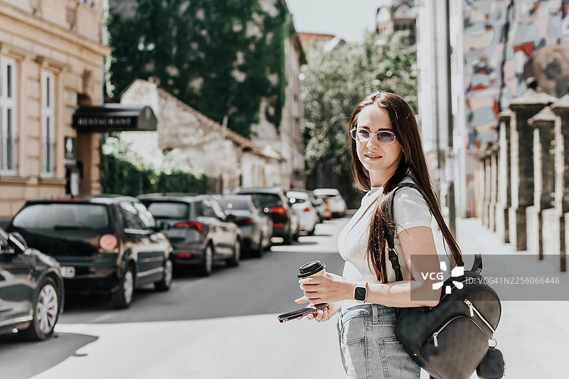 一位戴着太阳镜的年轻女子在城市街道上，一手拿着智能手机，一手拿着外卖咖啡。图片素材