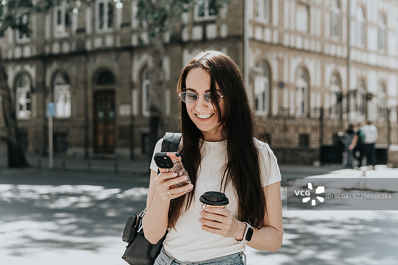 一位戴着太阳镜的年轻女子，脸上带着笑容，一手拿着智能手机，一手拿着外带咖啡，站在城市街道上。图片素材