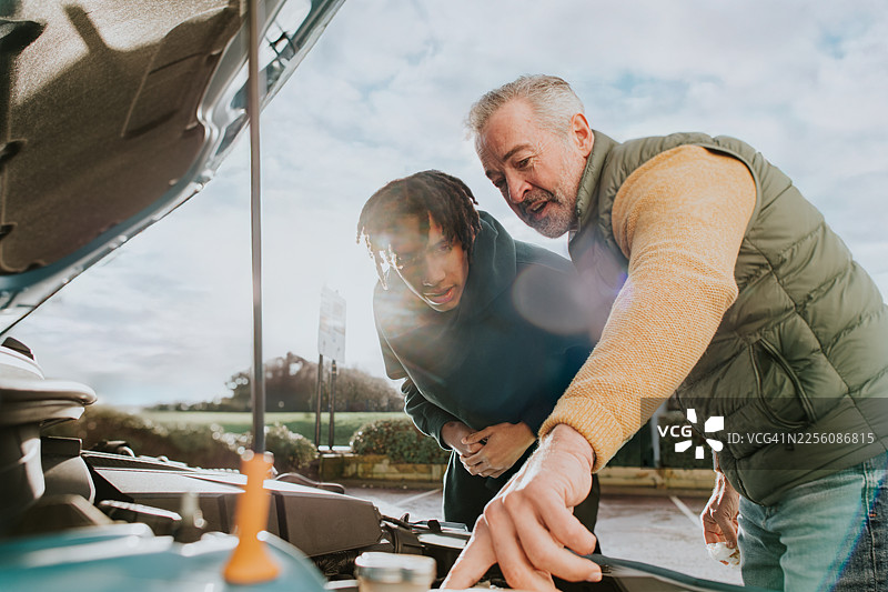 一位驾校教练向年轻男学员传授车辆维修知识，并指示了引擎盖下的水箱和机油。图片素材