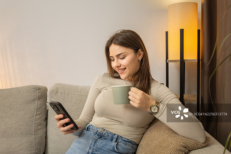 年轻女子躺在沙发上喝咖啡，同时查看手机图片素材