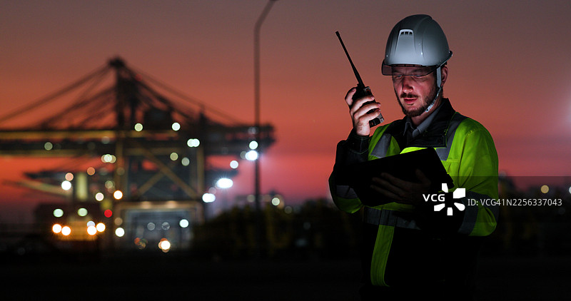 一名身穿防护装备的工业工人在日落时分于港口设施使用数字平板电脑，背景是起重机和货物基础设施。图片素材