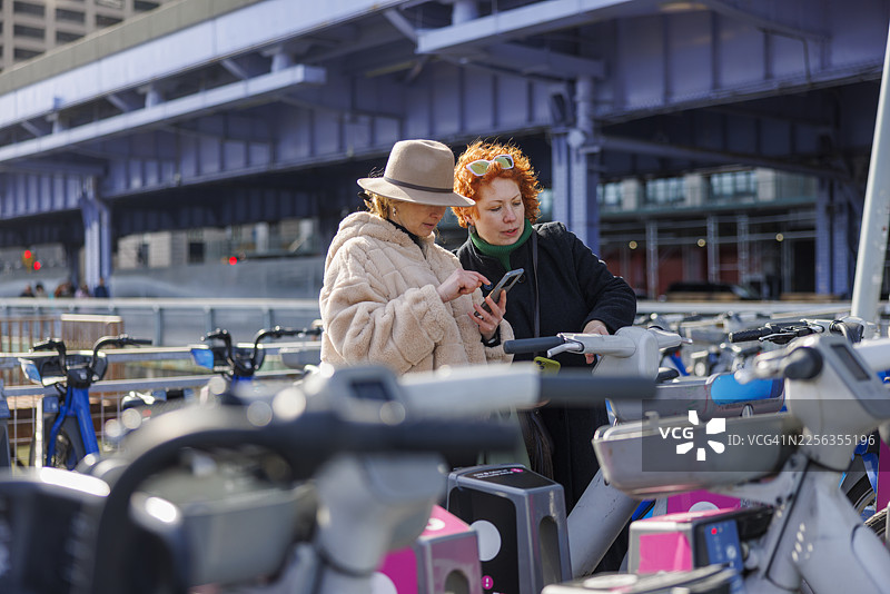 女性朋友们在曼哈顿使用智能手机应用程序预订共享单车服务。纽约市的城市旅游和可持续交通。图片素材