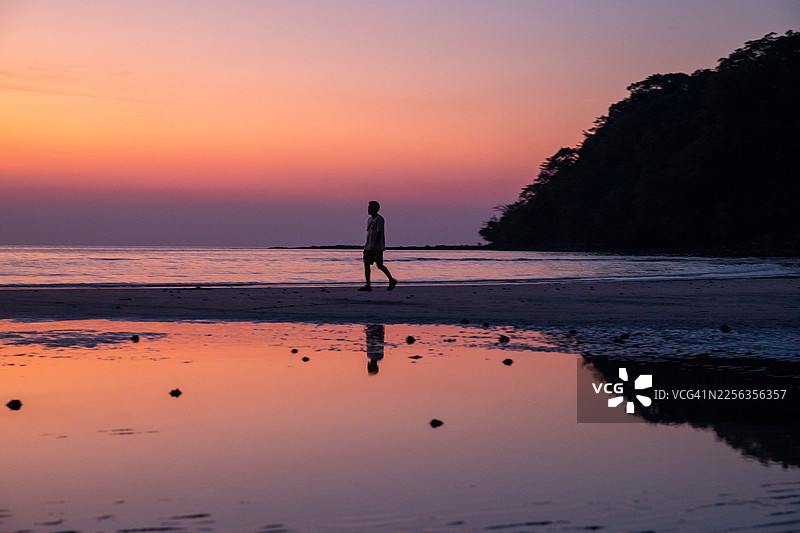 阁骨岛 - 黄昏时分，一名男子独自走在阁骨岛海岸线上的剪影。美丽的日落天空和在平静水面上的倒影。图片素材