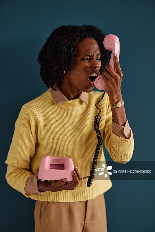 一位有色人种女性对着粉色复古电话大喊，背景是蓝色墙壁，表达着沮丧和沟通的能量图片素材