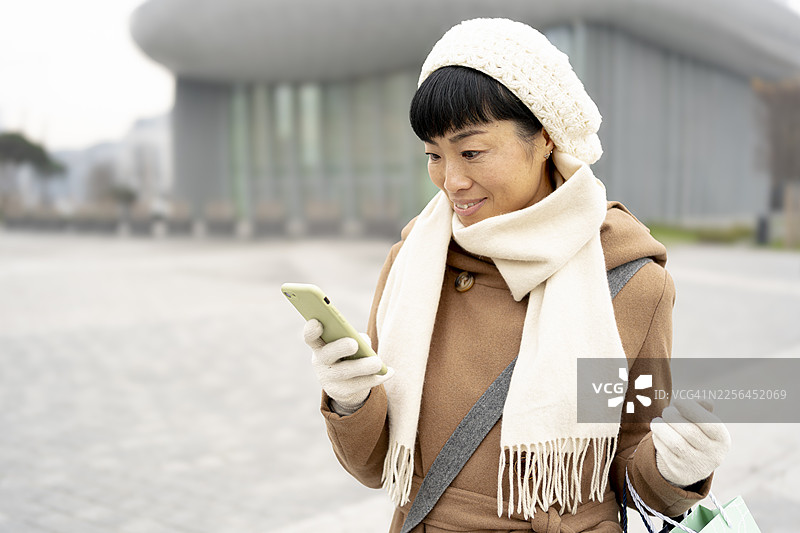 冬季城市里，一位亚洲女性微笑着使用智能手机图片素材