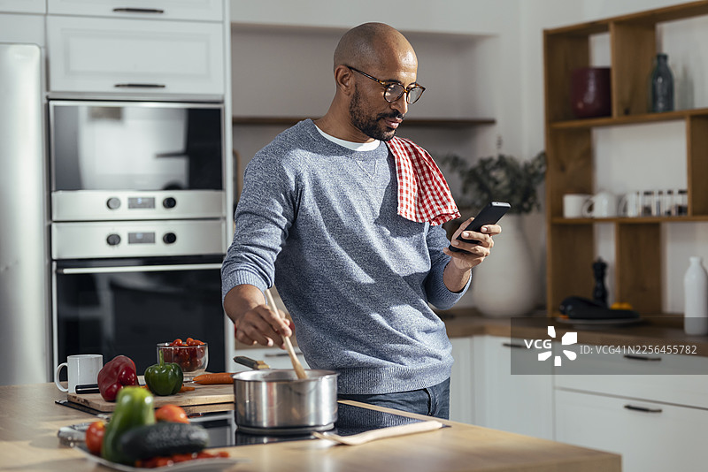 专注的男士在家中厨房使用智能手机并搅拌图片素材