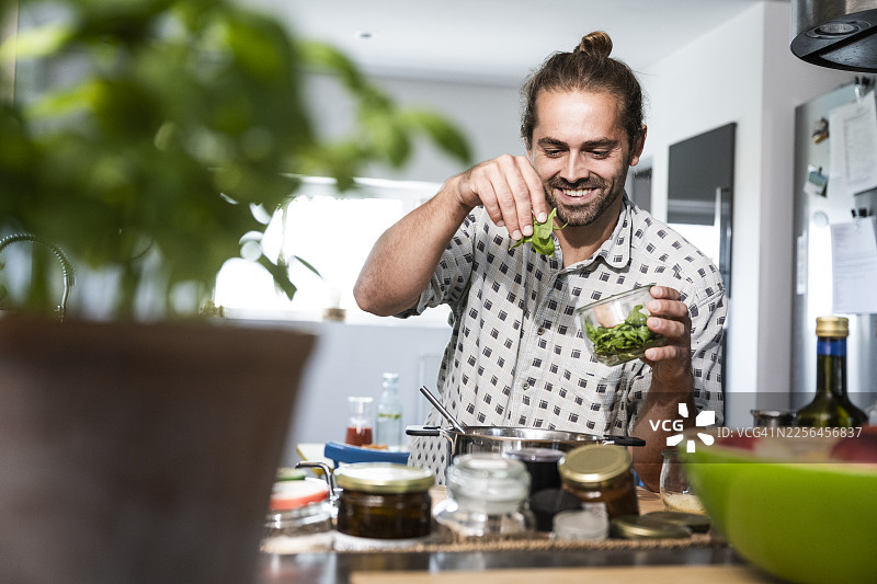 快乐的男人在家里的厨房里添加罗勒叶并烹饪食物图片素材