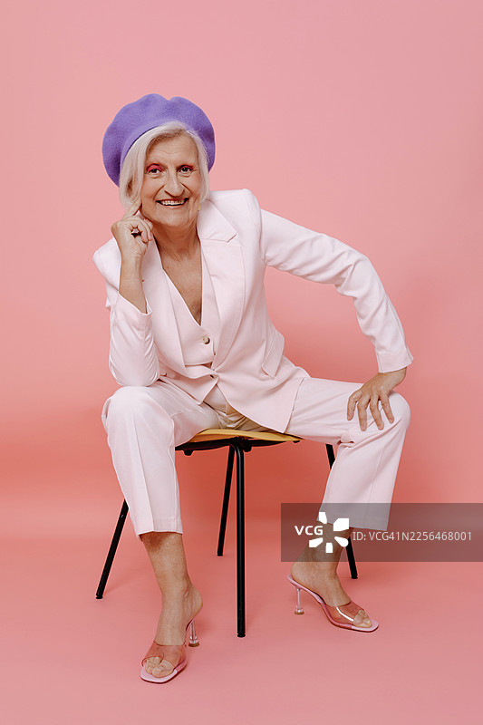 身穿白色西装、头戴紫色贝雷帽的快乐女子坐在粉色背景的椅子上图片素材