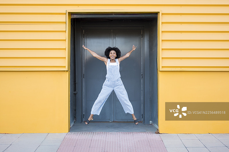 一位卷发非洲裔女性在黄色墙壁前快乐地跳跃图片素材