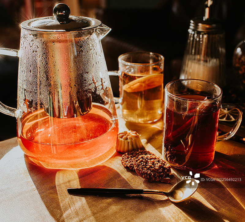 阳光下的暖色自然光，照亮了摆放着茶点和两杯茶的玻璃茶壶，置于桌上。图片素材