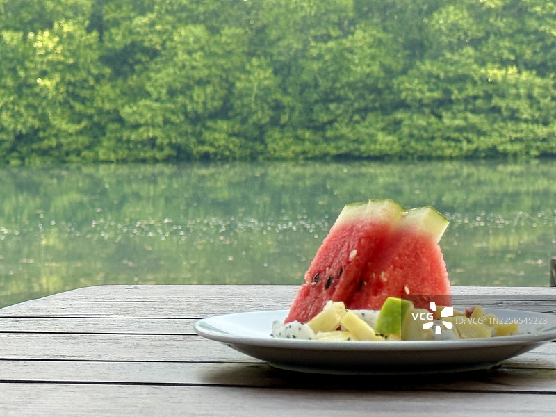 戈公岛：在宁静河水中，木桌上摆放着一份新鲜水果早餐，背景是生机勃勃的绿色红树林及其倒影。图片素材