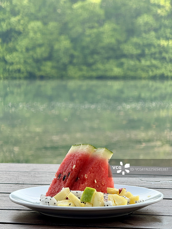 戈公岛：在宁静河水中，以生机勃勃的绿色红树林和倒影为背景，木桌上摆放着一份新鲜水果早餐。图片素材
