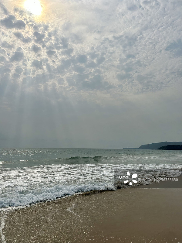 阳光穿透云层，照射在宁静的海洋和沙滩上，远处是印度南果阿的山丘图片素材