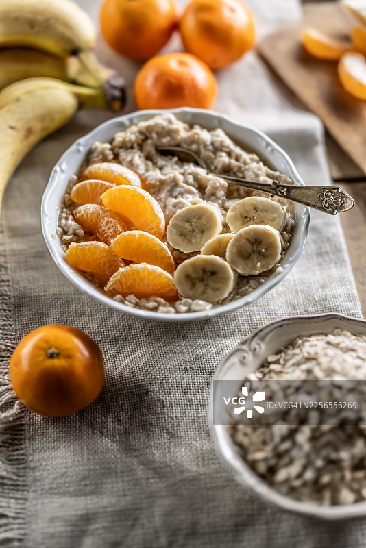 特写一碗温暖的燕麦粥，上面装饰着香蕉片和橘子瓣，淋着枫糖浆，摆放在乡村风格的餐桌上，背景有新鲜水果。图片素材