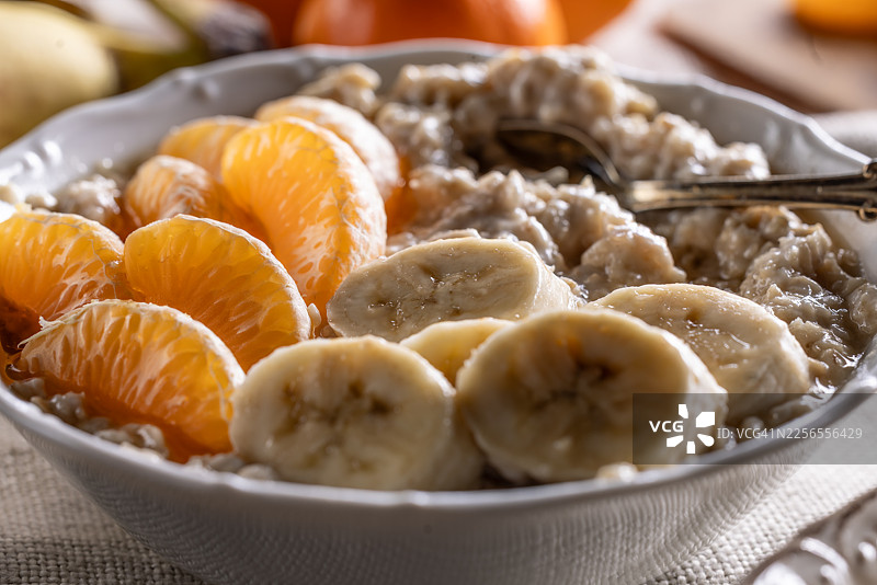 近景特写一碗温暖的燕麦粥，上面装饰着香蕉片和橘子块，淋着枫糖浆，摆放在一张乡村风格的桌子上，背景是新鲜的水果。图片素材