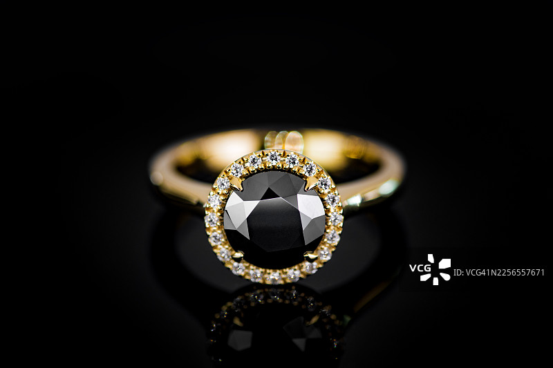 在反光的黑色背景上，一枚镶嵌着黑色钻石并被小白钻环绕的金色戒指的优雅特写图片素材