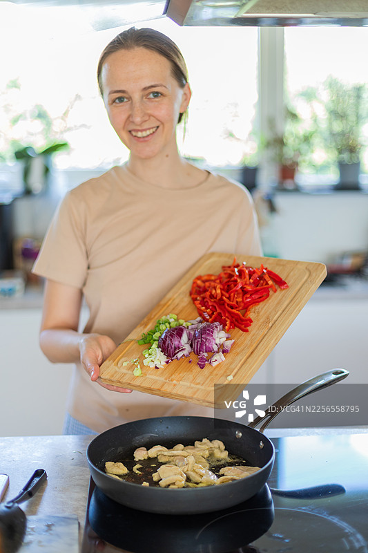 一位女士微笑着在现代厨房里准备健康餐食，往煎锅里添加新鲜蔬菜图片素材