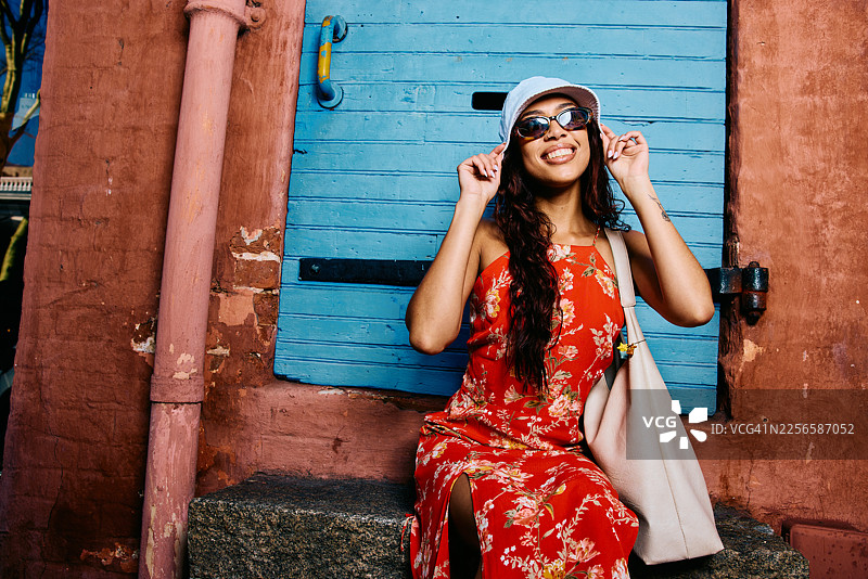 一位二十多岁的年轻旅行者，身穿色彩鲜艳的夏日连衣裙，倚靠在一堵充满活力的蓝色墙壁旁，脸上带着微笑。图片素材