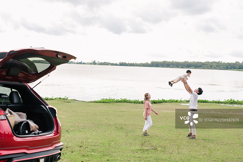 亚洲一家三口在湖边享受公路旅行。父亲开心地将蹒跚学步的儿子举起，母亲在一旁微笑，享受亲子时光、欢声笑语、自由和旅行目的地。图片素材