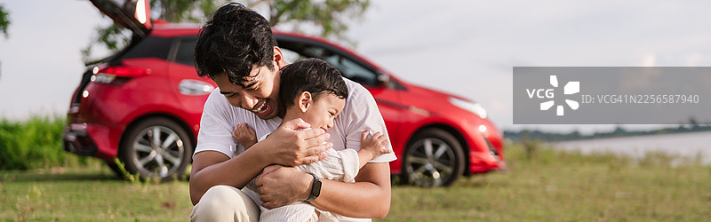 亚洲父亲在湖边拥抱他微笑的幼儿儿子。家庭在公路旅行中增进感情，家庭旅行度假。交通，旅行保险概念图片素材