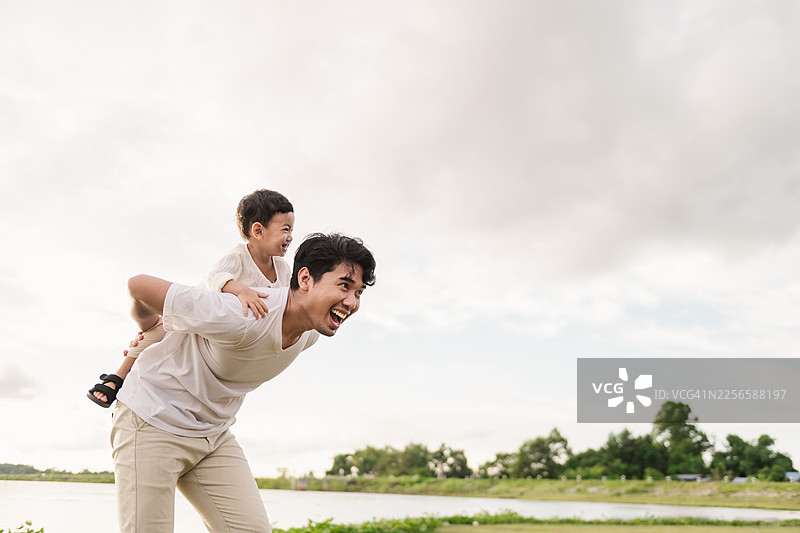父亲时间与儿子在户外自然公园玩耍，亚洲父亲背着蹒跚学步的孩子进行户外探险，展现了爱、情感、亲情、享受、无忧无虑的周末活动图片素材