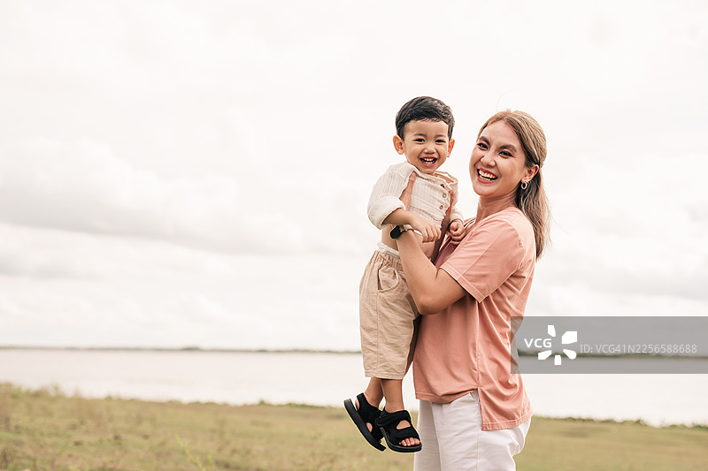 亚洲母亲抱着蹒跚学步的儿子在湖边微笑。一家三口享受亲子时光，充满爱意、幸福和无忧无虑的生活方式，适合夏日周末户外活动、自然公园，展现积极情绪。图片素材