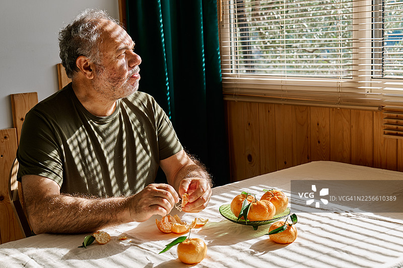 一位成熟的男士坐在桌前，吃着橘子并望着窗外。图片素材