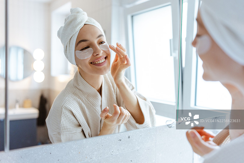 年轻女子在浴室里享受早晨的自我护理，脸上贴着眼膜，身穿睡袍。图片素材