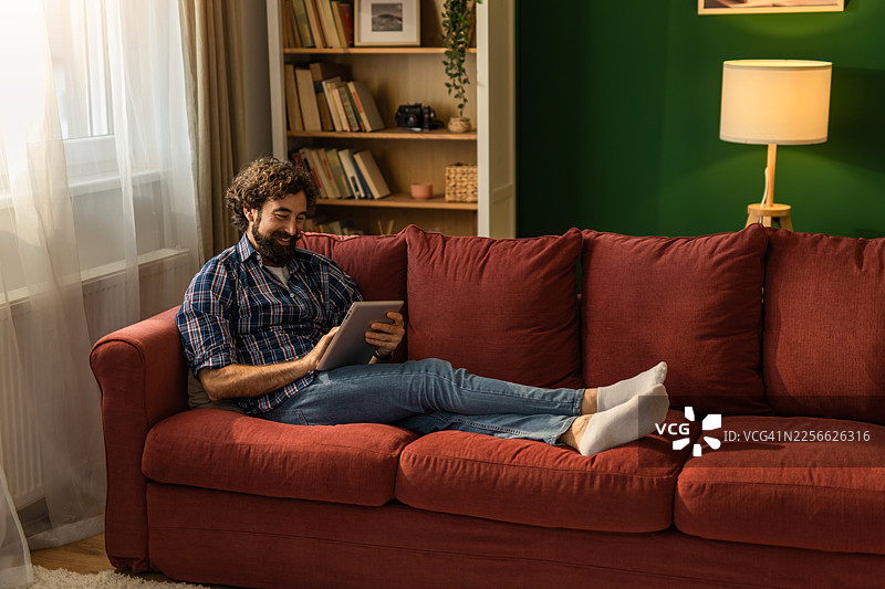 男人放松地躺在沙发上，微笑着使用平板电脑图片素材