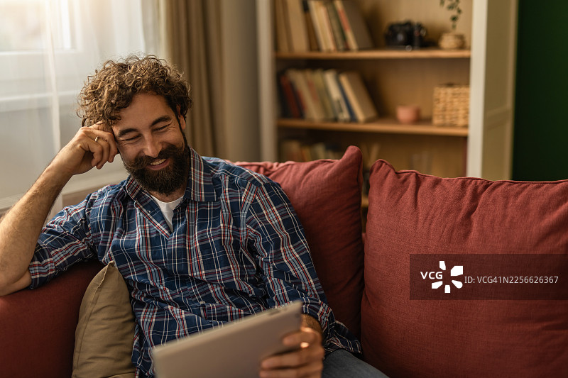 年轻男子躺在沙发上，一手拿着平板电脑，一手扶着头，开心地大笑图片素材
