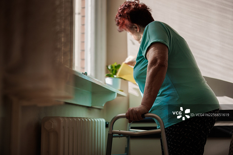 一位年长的妇女拄着助行器在窗边给植物浇水，象征着关爱与独立图片素材